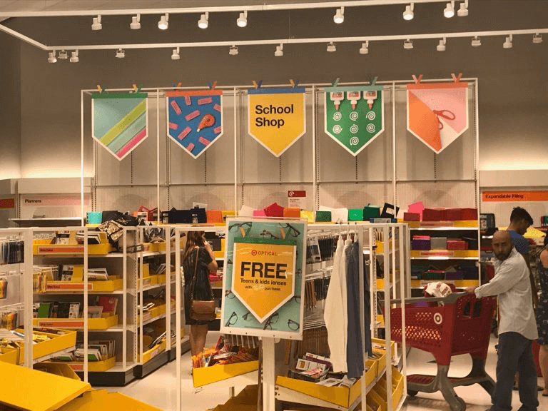 Retail marketing: A store section decorated with colorful banners labeled “School Shop”, displaying shelves filled with school supplies, such as notebooks and folders. Shoppers browse the items and a sign advertises free kids lenses near the entrance.